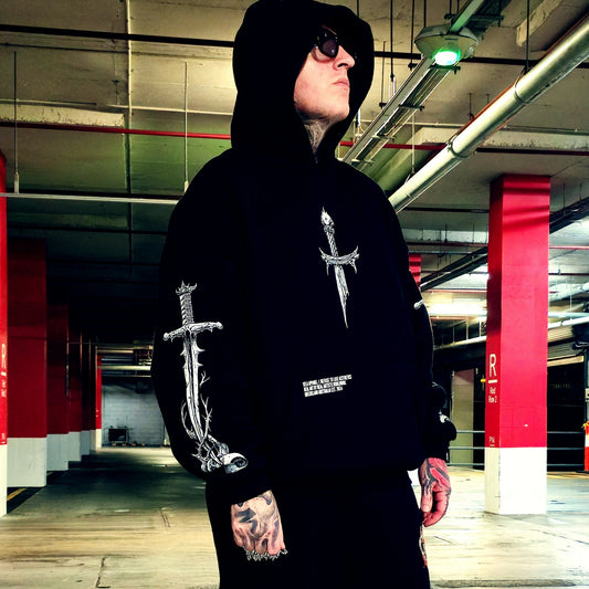 A person wearing a black hooded sweatshirt with white graphic sword illustrations on the chest and sleeve, standing in a concrete parking garage.