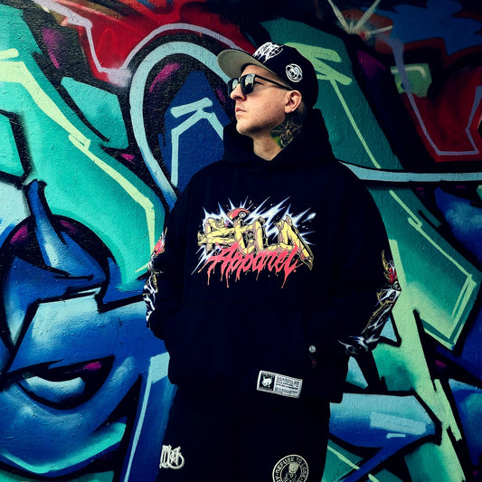 Person wearing a black hoodie with colourful text in front of a graffiti wall. RTLA-Apparel