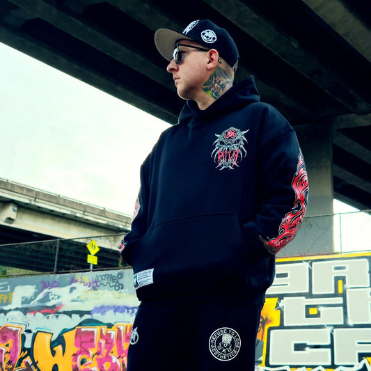 Person wearing a black hoodie with a colourful design, standing under an overpass with graffiti in the background. RTLA-Apparel