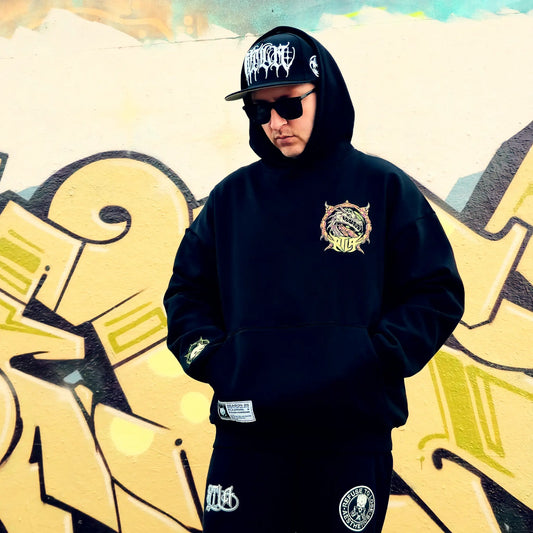 Person wearing a black hoodie with a logo, sunglasses, and a cap in front of graffiti art. RTLA-Apparel