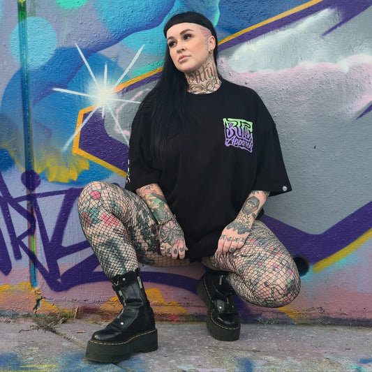 Person with tattoos crouching in front of a colorful graffiti wall