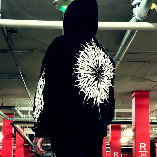 A person viewed from behind wearing a black hoodie with large white graphic prints on the back and sleeves, standing in an indoor parking structure with red support columns visible in the background.