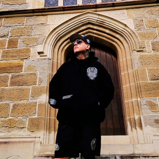 Person wearing a black hoodie with a logo in front of a stone building. RTLA-Apparel
