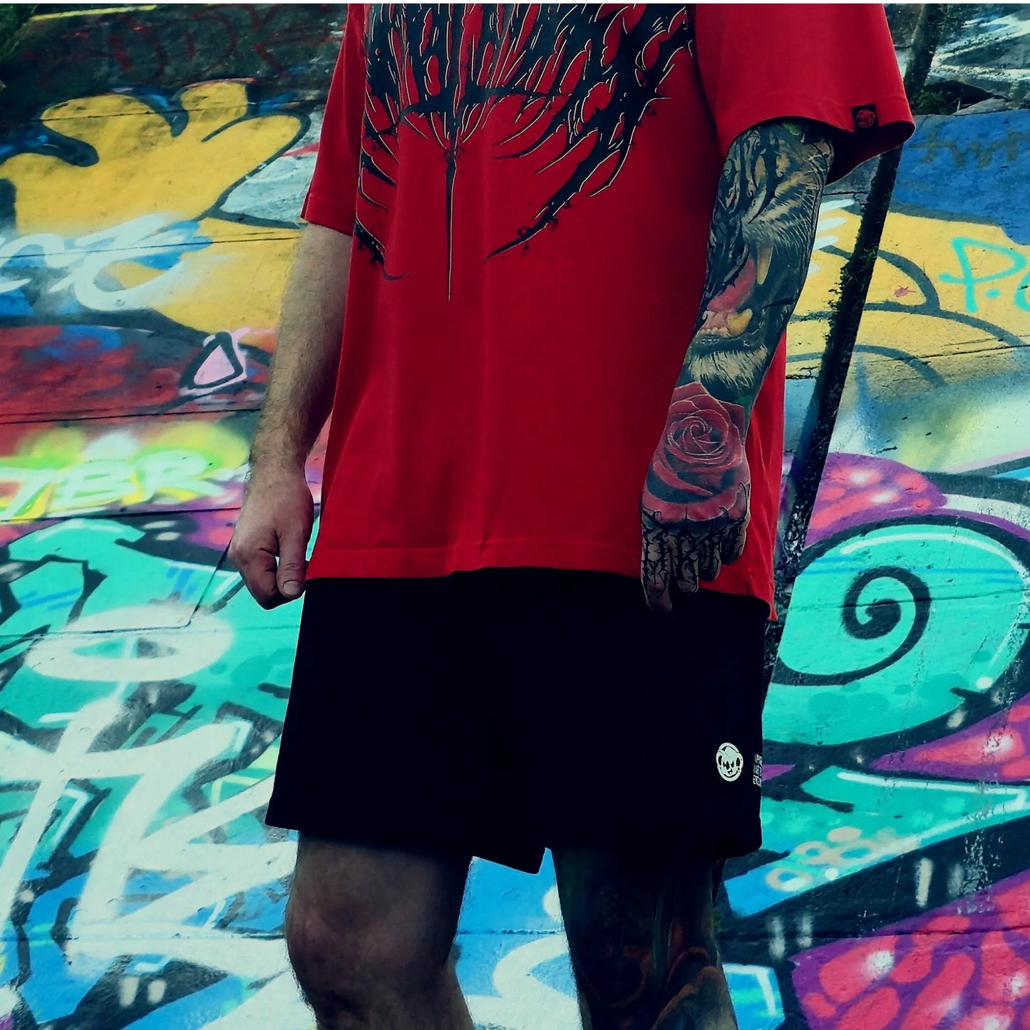 Person wearing a red shirt and black shorts with tattoos, standing against a colorful graffiti background.