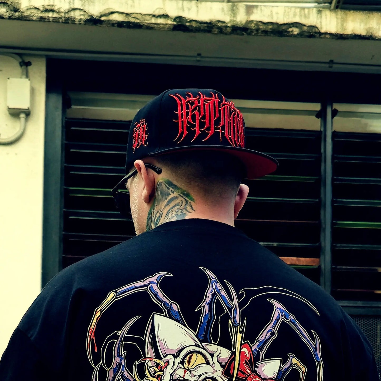 Person wearing a black cap with red text and a black shirt with a colorful design, standing in front of a building.