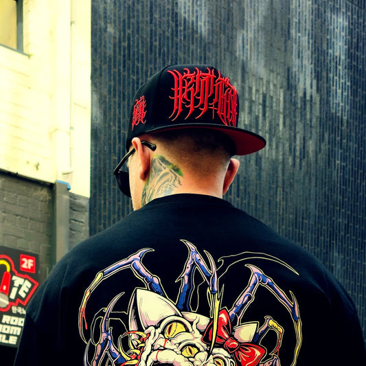 Person wearing a black cap with red designs and a black shirt with a colorful graphic, standing against a textured wall.