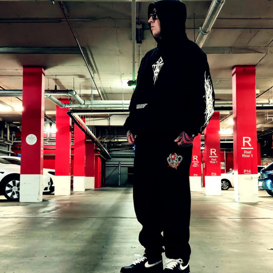 A person wearing a black hooded sweatshirt and matching black sweatpants stands in a parking garage. The outfit has white graphic prints on the chest and sleeve, a small heart patch on the thigh, and black-and-white sneakers.