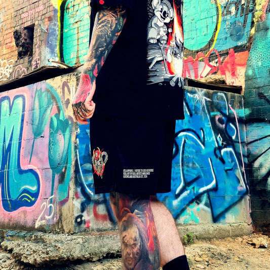 A person wearing a black graphic t-shirt and matching black shorts with white text and a cartoon print, standing in front of a graffiti-covered brick wall.