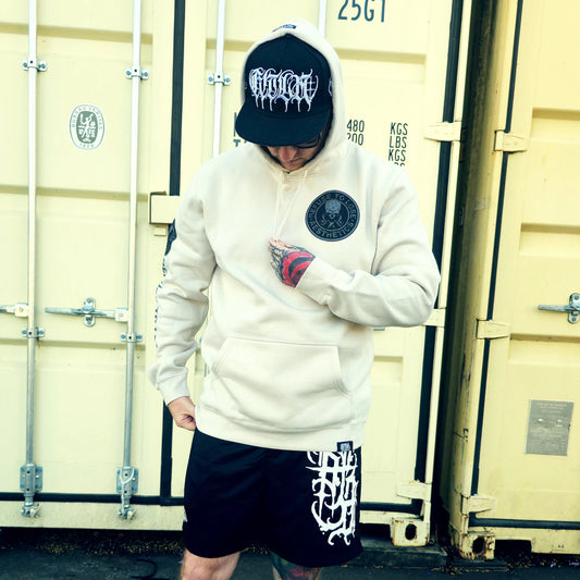 Person wearing a white hoodie and black cap with a logo, standing in front of shipping containers.