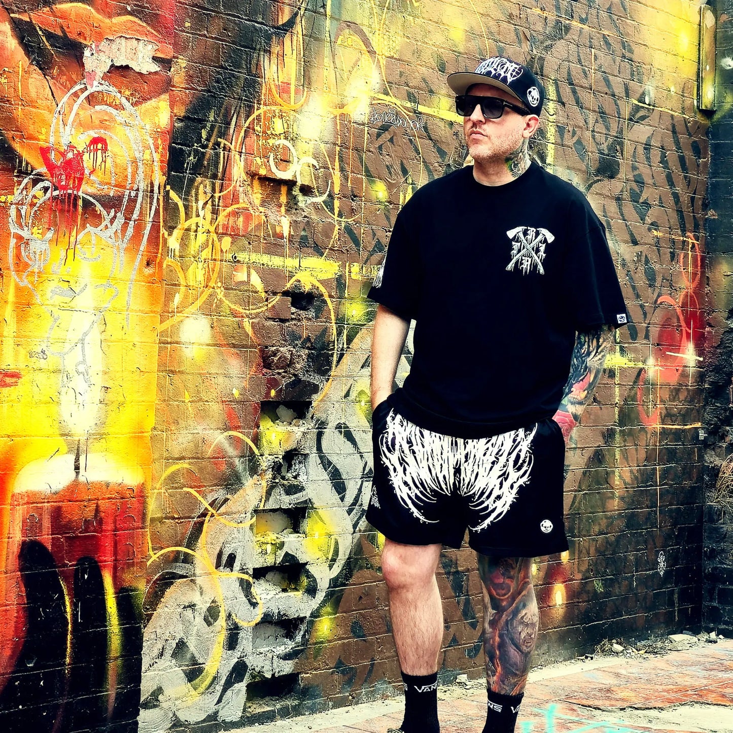 Person wearing a black t-shirt and shorts with white designs, standing in front of a colorful graffiti wall.