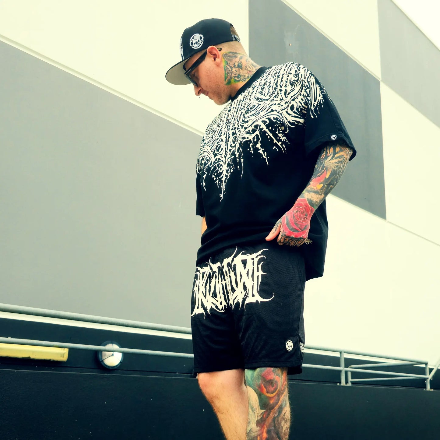 Man wearing a black graphic t-shirt and shorts with tattoos, standing outdoors.
