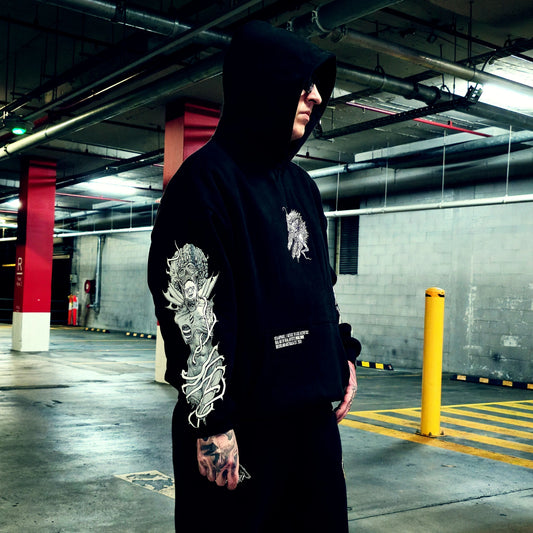 A person wearing a black hooded sweatshirt with detailed white graphic prints on the sleeve and chest, standing in a concrete parking garage.