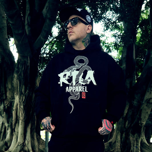 Person wearing a black hoodie with a logo and text, standing in a forest setting.