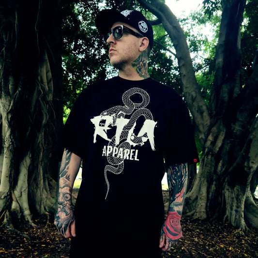 Person wearing a black t-shirt with a white and red graphic design of a mask and sword, standing in a natural setting.