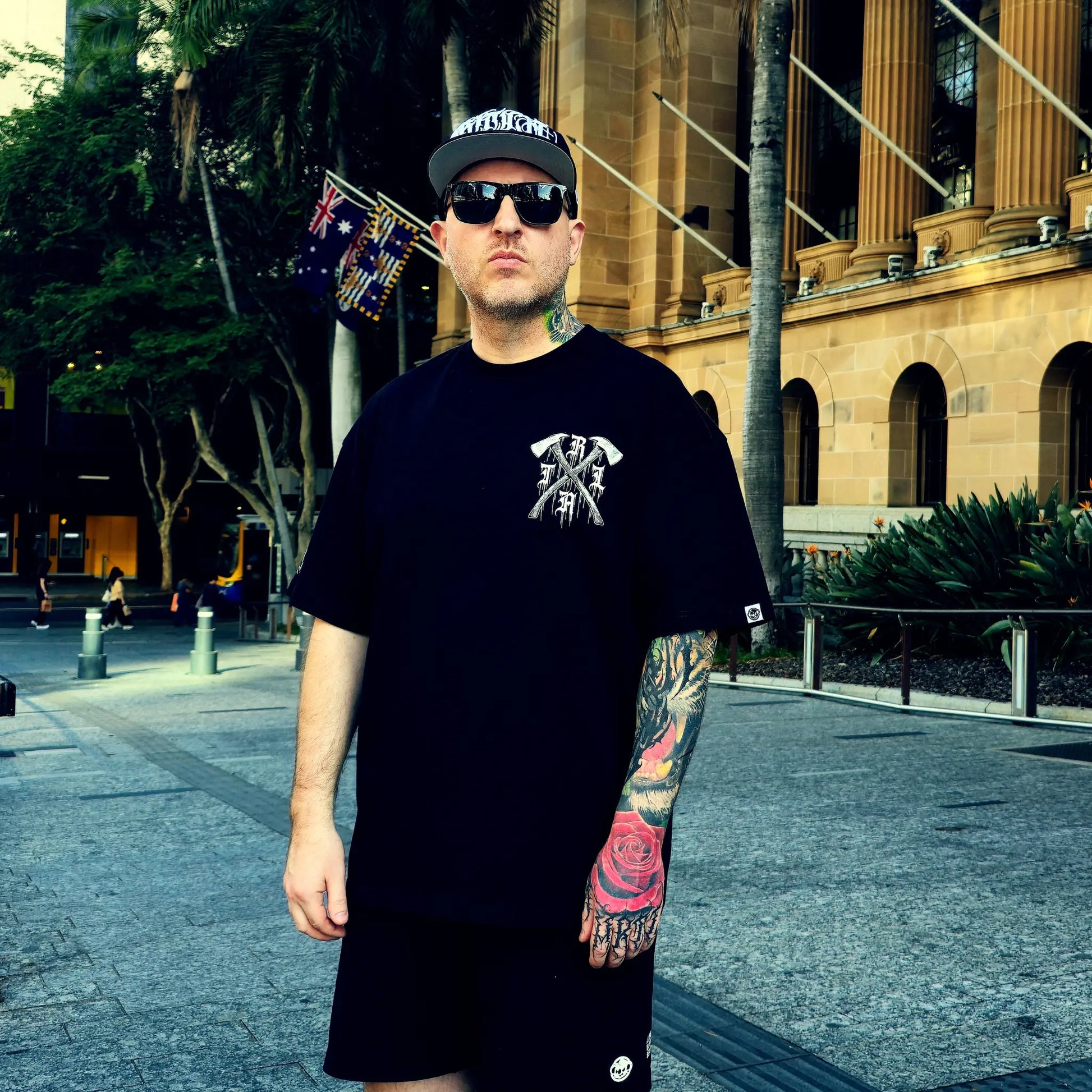 Man wearing a black t-shirt with a logo and sunglasses, standing in an urban setting.