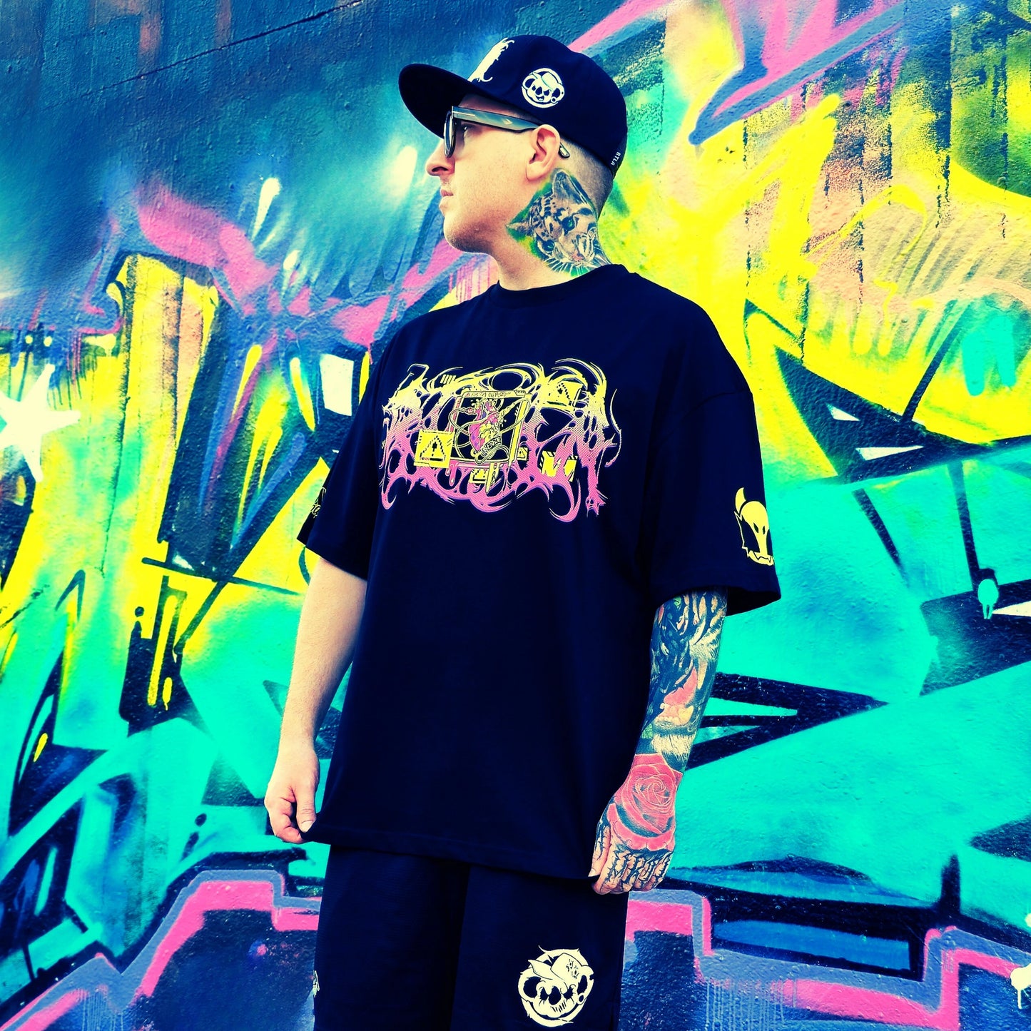 Person wearing a black t-shirt with colorful graphics in front of a vibrant graffiti wall.