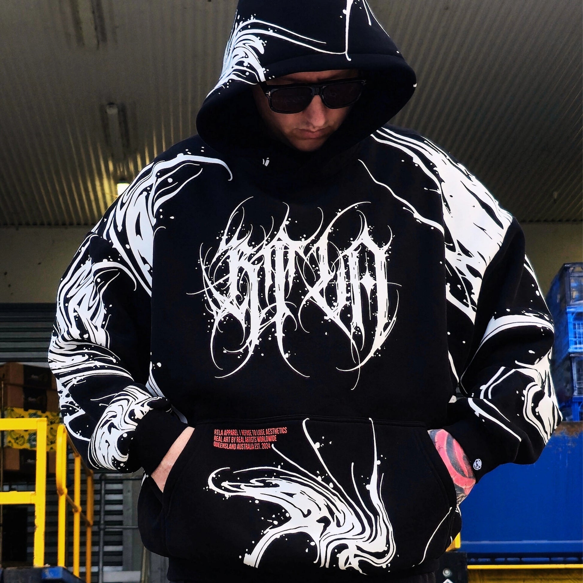Person wearing a black hoodie with white designs and text, standing in an industrial setting.
