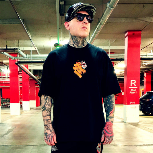 A tattooed man wearing a black t-shirt with a yellow and white graphic, a black cap, and sunglasses, standing in a parking garage.