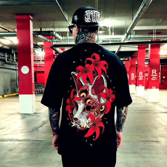 A man viewed from behind wearing a black t-shirt with a large red and white graphic of a fox surrounded by red flowers on the back, standing in a concrete parking garage with red pillars.