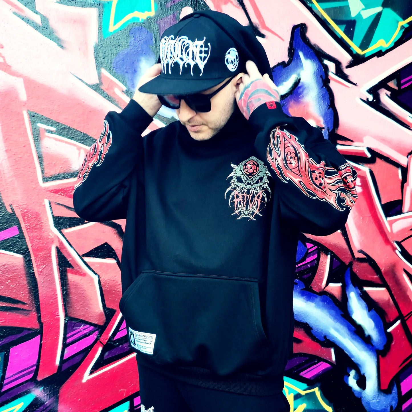 Person wearing a black hoodie with a floral design and a cap in front of colourful graffiti. RTLA-Apparel