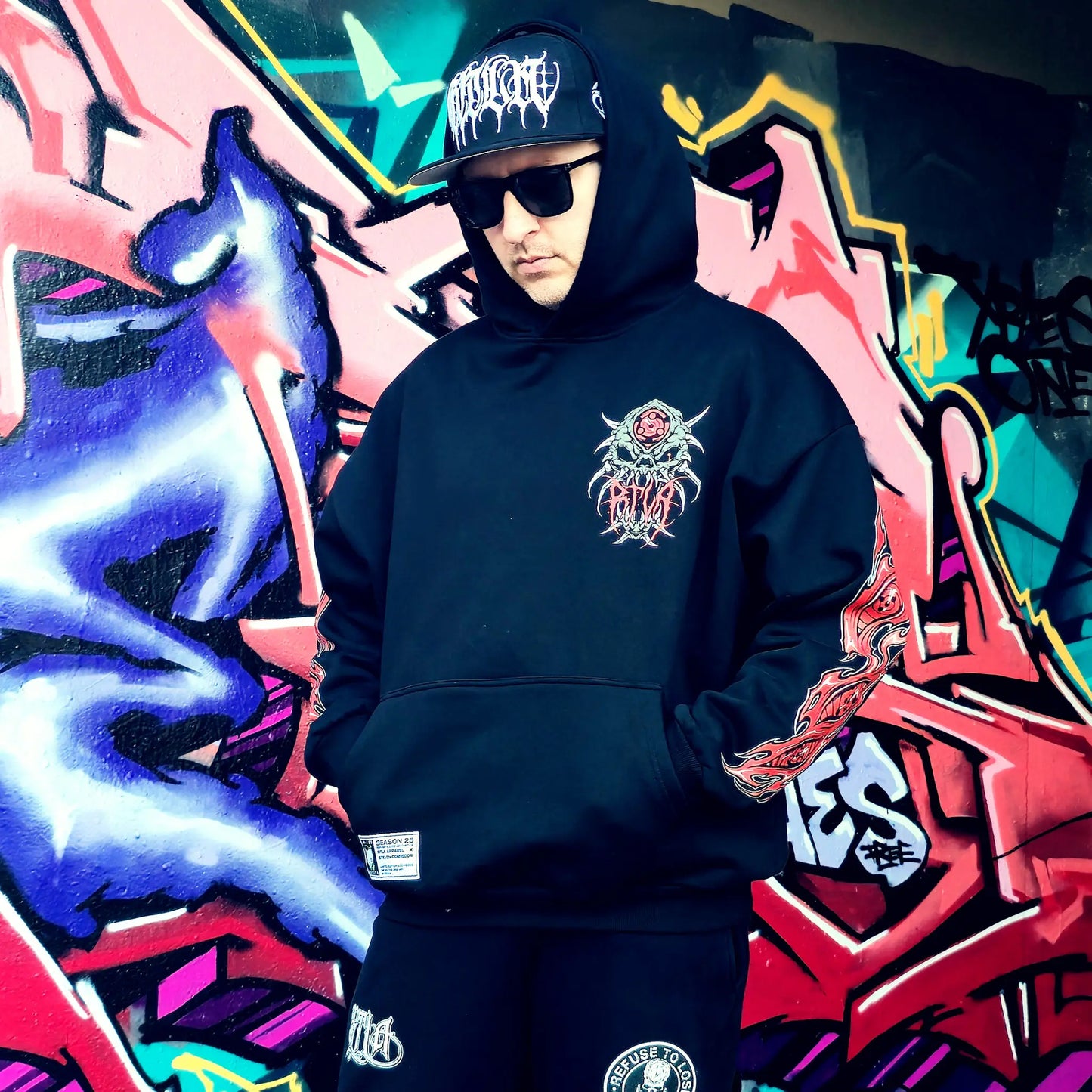 Person wearing a black hoodie with a logo in front of colourful graffiti art. RTLA-Apparel