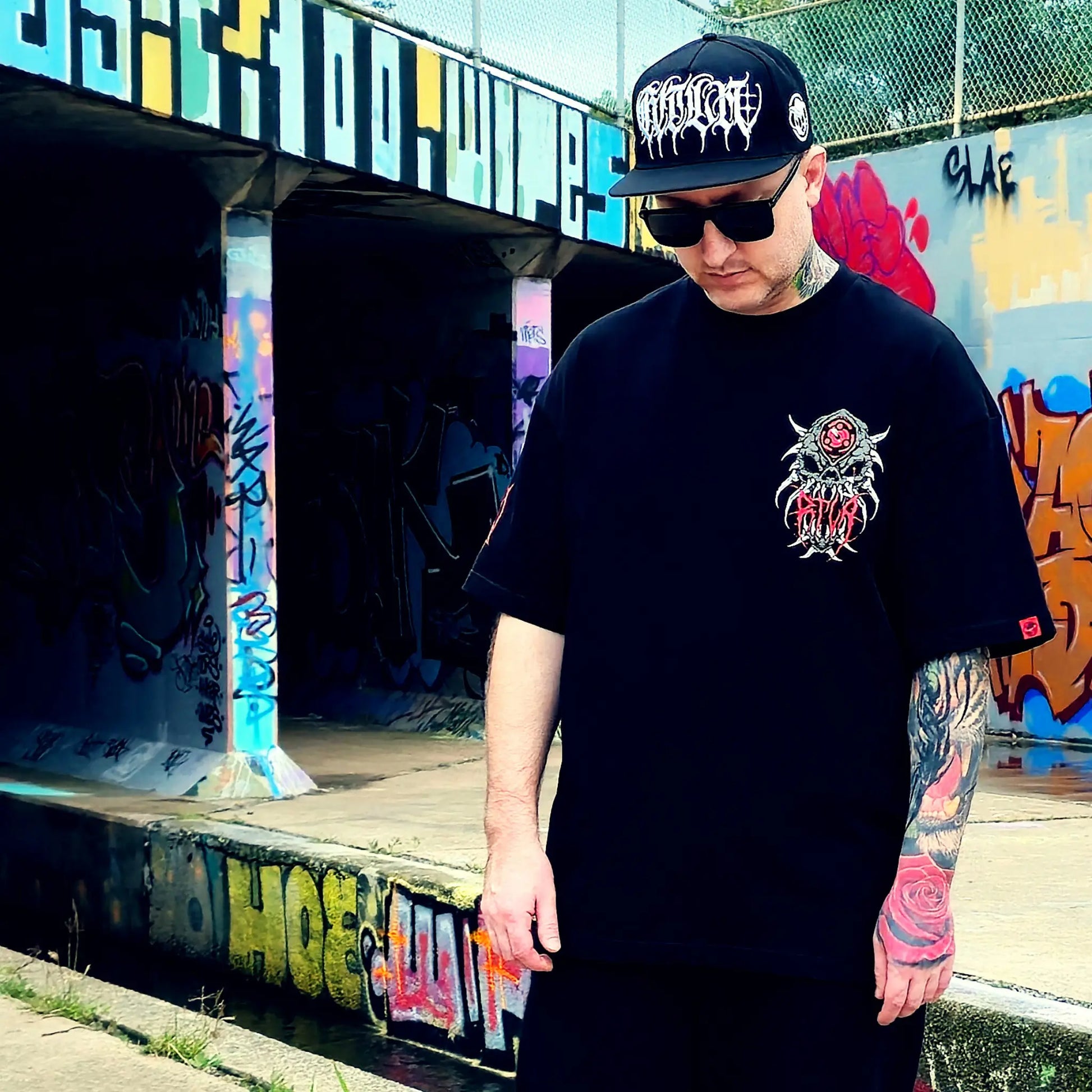 Person wearing a black t-shirt with a graphic design and a cap, standing in front of graffiti-covered walls.