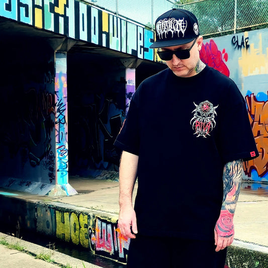 Person wearing a black t-shirt with a graphic design and a cap, standing in front of graffiti-covered walls.