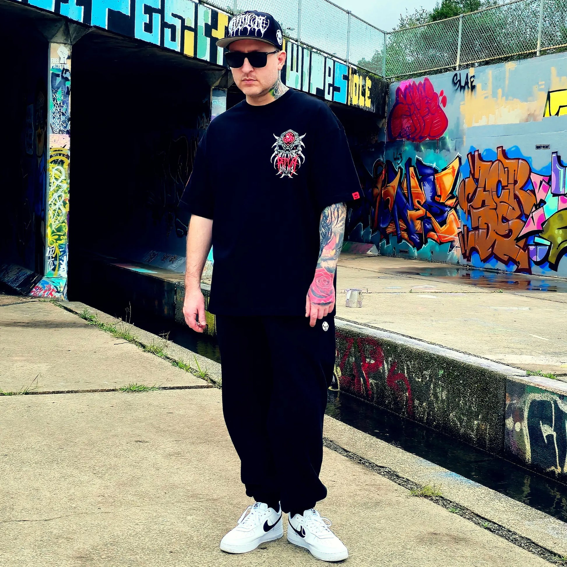 Person wearing a black t-shirt with a logo and text, black pants, and white sneakers, standing in front of a graffiti-covered wall.