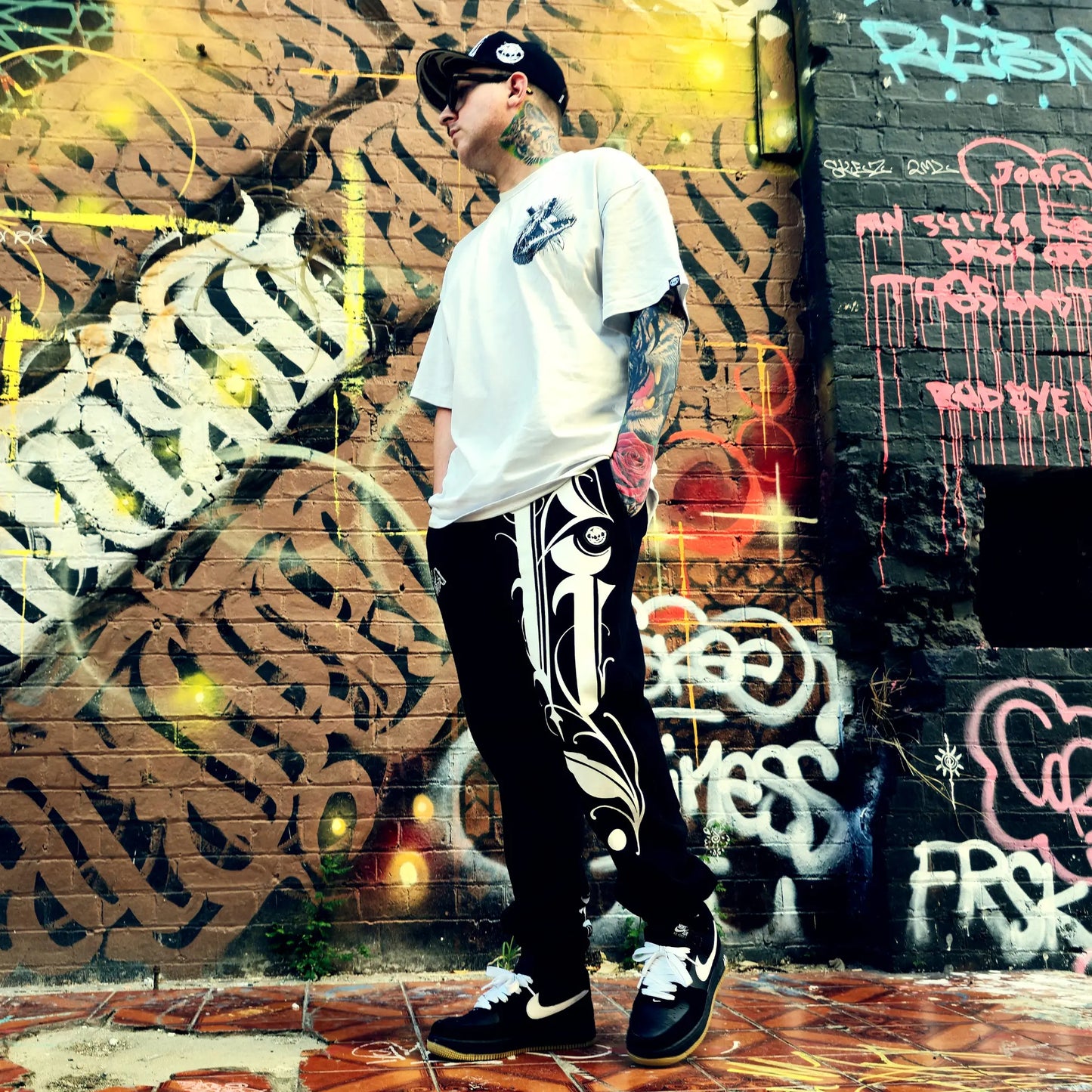 Person wearing a white t-shirt and black pants with white patterns, standing in front of a graffiti-covered wall.