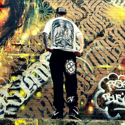 Person wearing a white shirt with a graphic design and black pants standing in front of a colorful graffiti wall.
