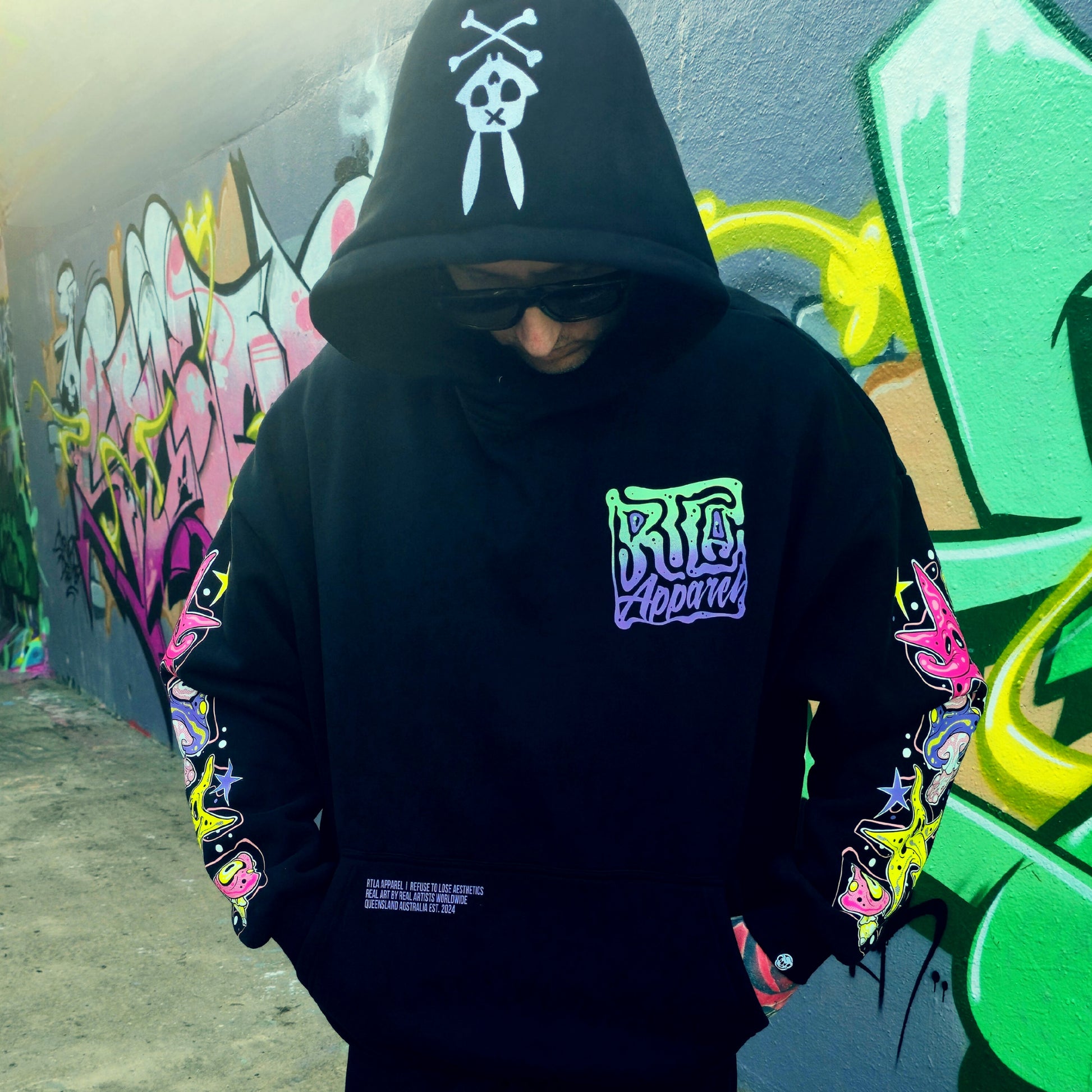 Person wearing a black hoodie with colorful designs and text, standing in front of a graffiti-covered wall.