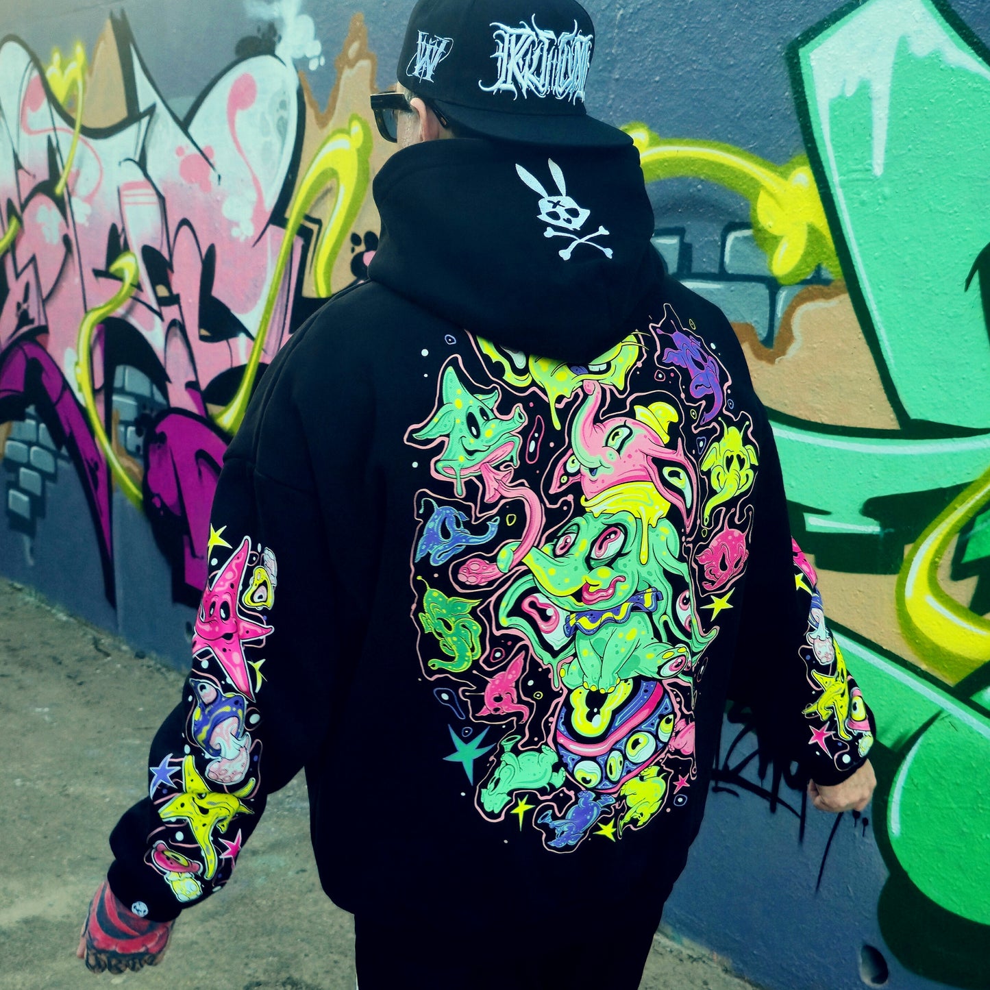 Person wearing a black hoodie with colorful designs in front of a graffiti-covered wall.