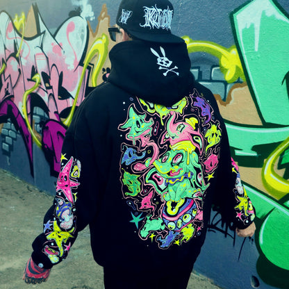Person wearing a black hoodie with colorful designs in front of a graffiti-covered wall.