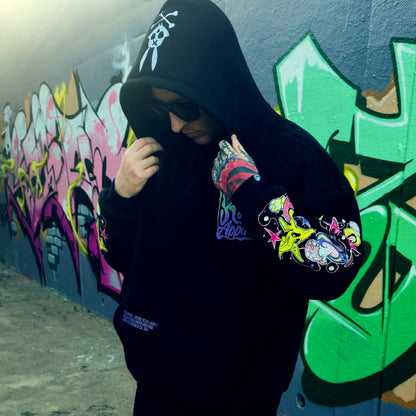 Person wearing a black hoodie with colorful designs in front of a graffiti-covered wall.
