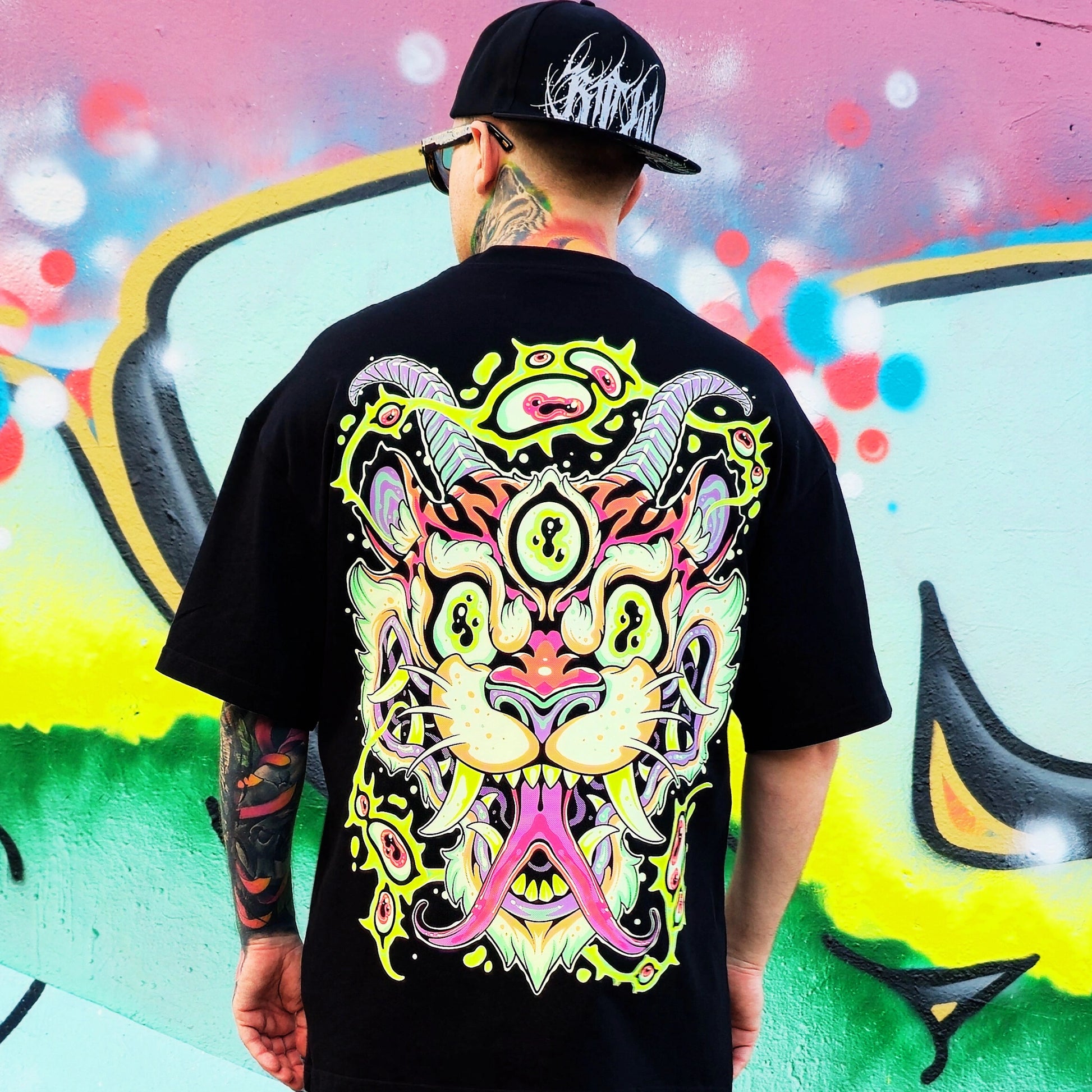 Person wearing a black t-shirt with a colorful graphic design, standing in front of a vibrant graffiti wall.