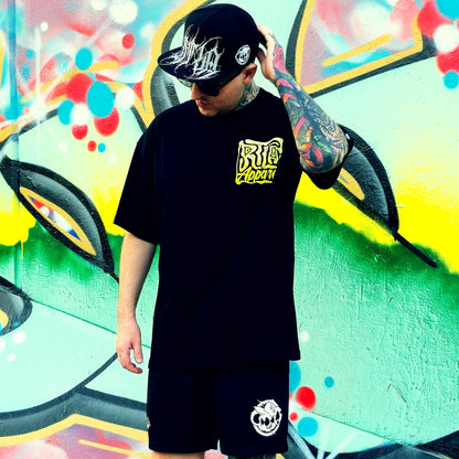 Person wearing a black t-shirt and shorts with logos, standing in front of a colorful graffiti wall.