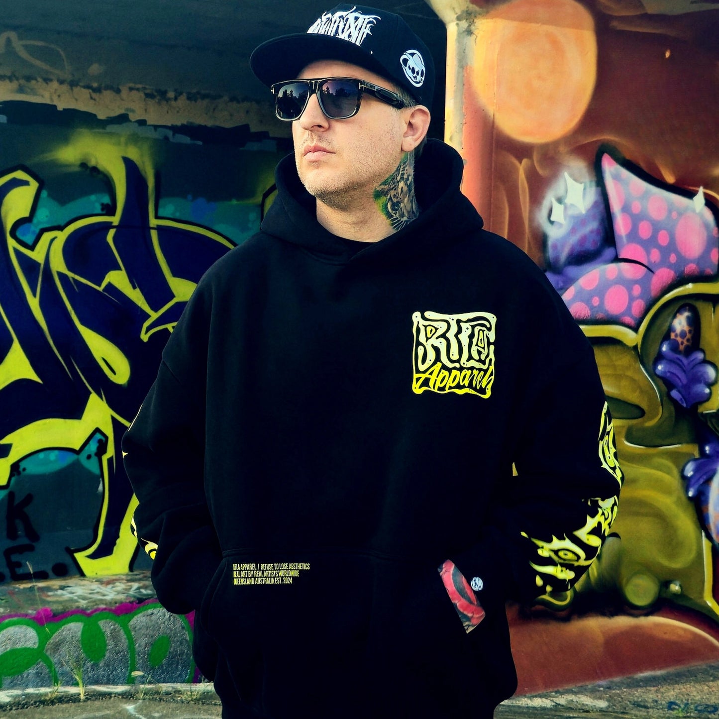 Man wearing a black hoodie with yellow text and graphics, sunglasses, and a cap in front of a graffiti wall.