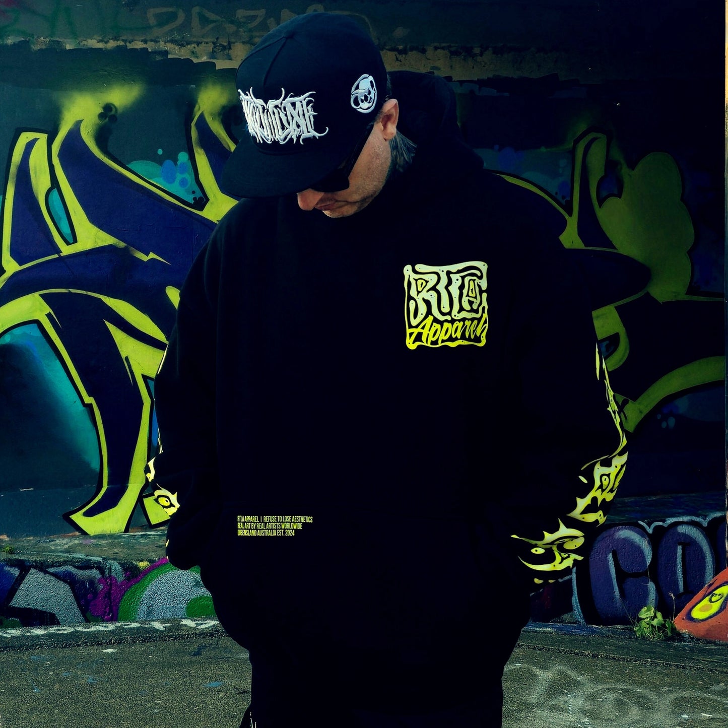 Person wearing a black hoodie with yellow text and a black cap with white text, standing in front of graffiti art.