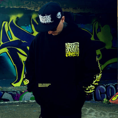 Person wearing a black hoodie with yellow text and a black cap with white text, standing in front of graffiti art.