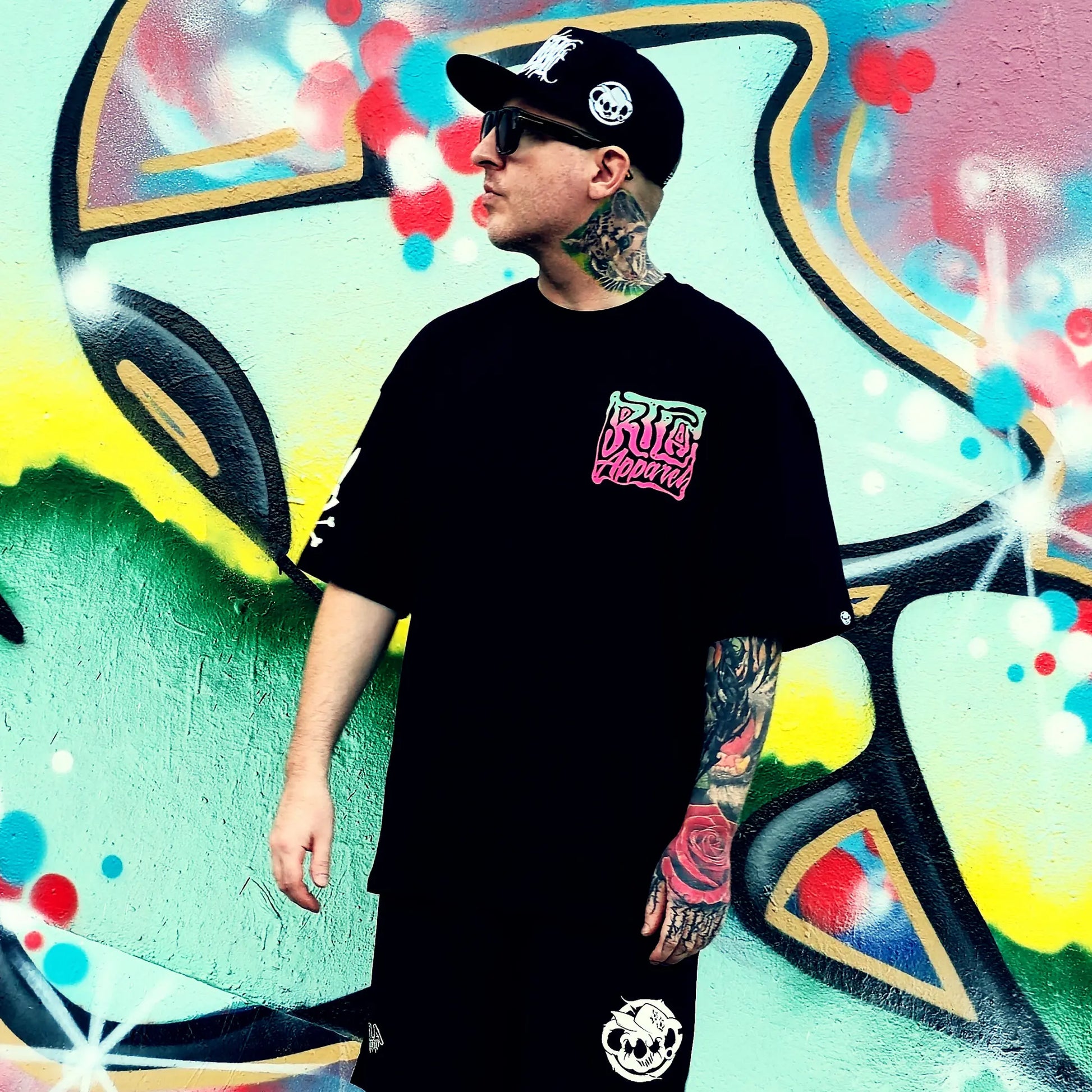 Person wearing a black t-shirt with colorful text, standing in front of a vibrant graffiti wall.