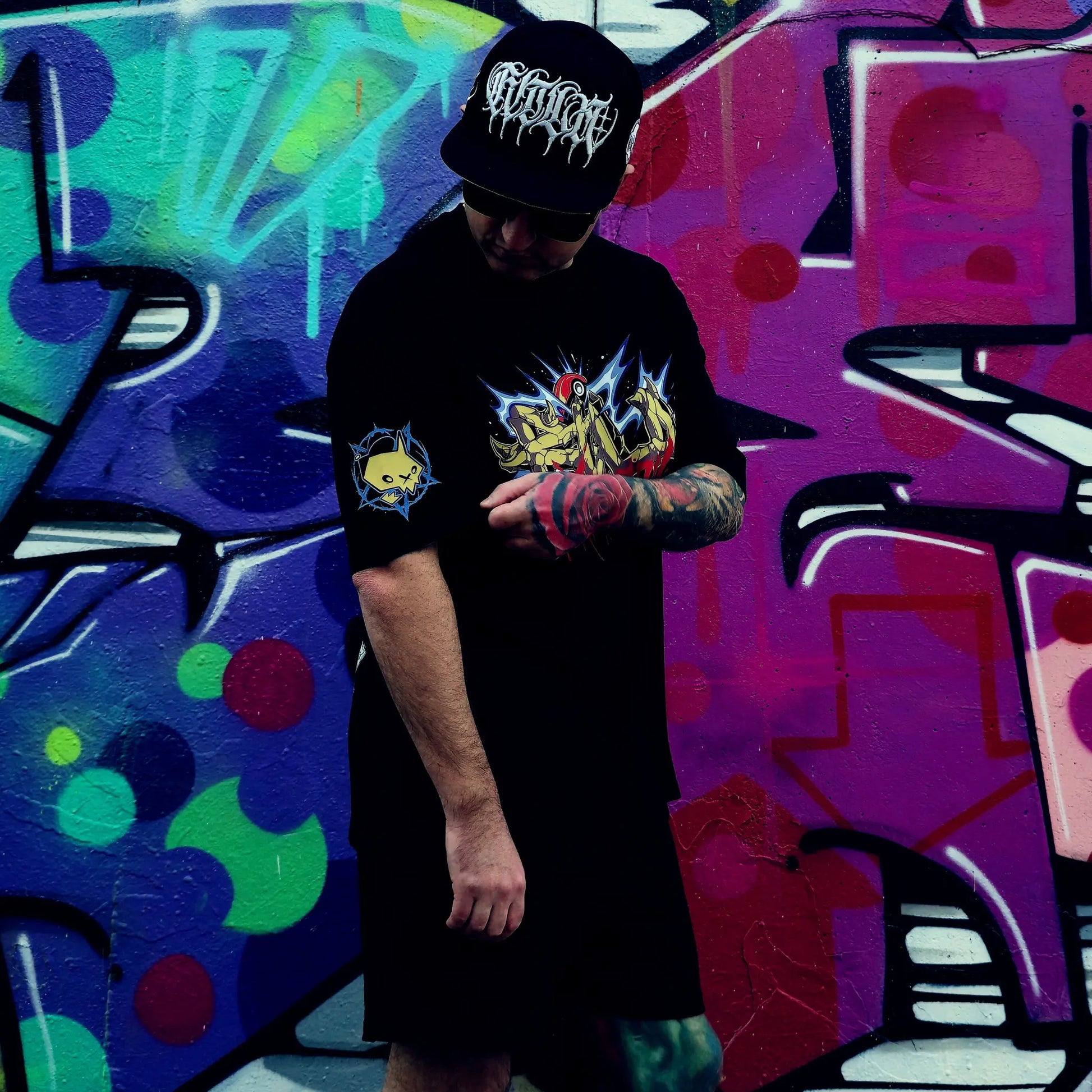 Person wearing a black t-shirt with a graphic design and a cap, standing in front of a colourful graffiti wall. RTLA-Apparel