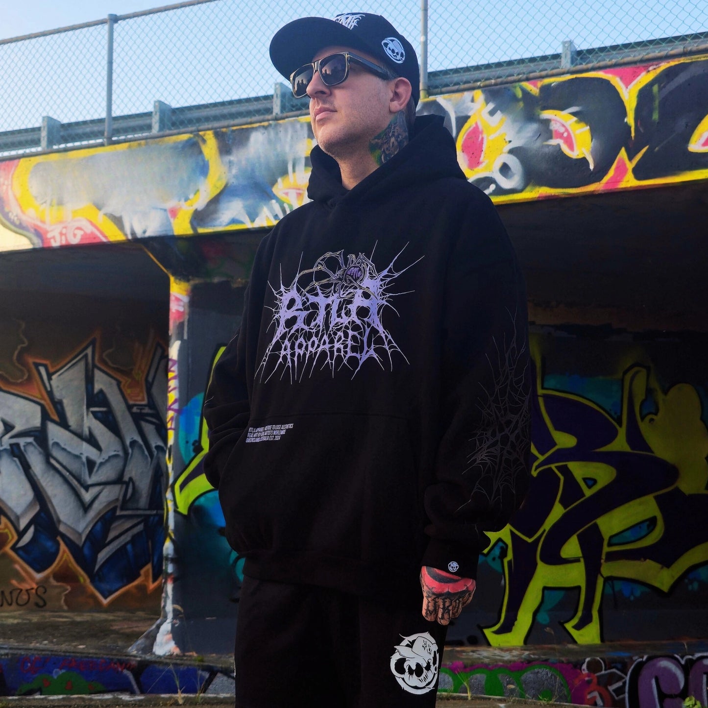 Person wearing a black hoodie with graphic design in front of colorful graffiti