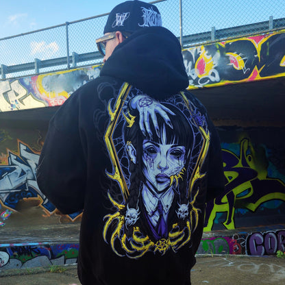 Person wearing a black hoodie with a graphic design of a woman's face and skulls, standing in front of a graffiti-covered wall.