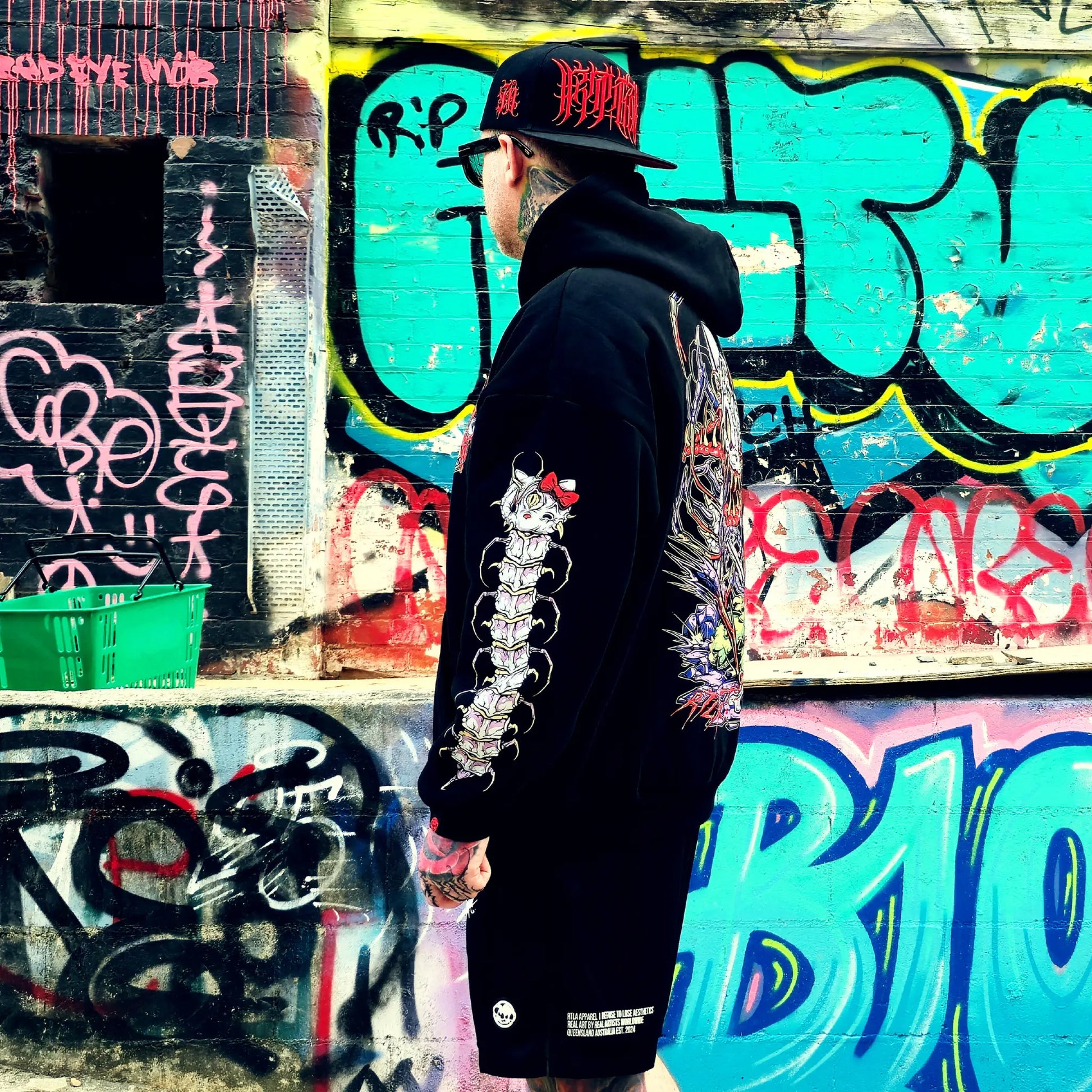Person wearing a black hoodie with a unique design in front of a graffiti-covered wall.