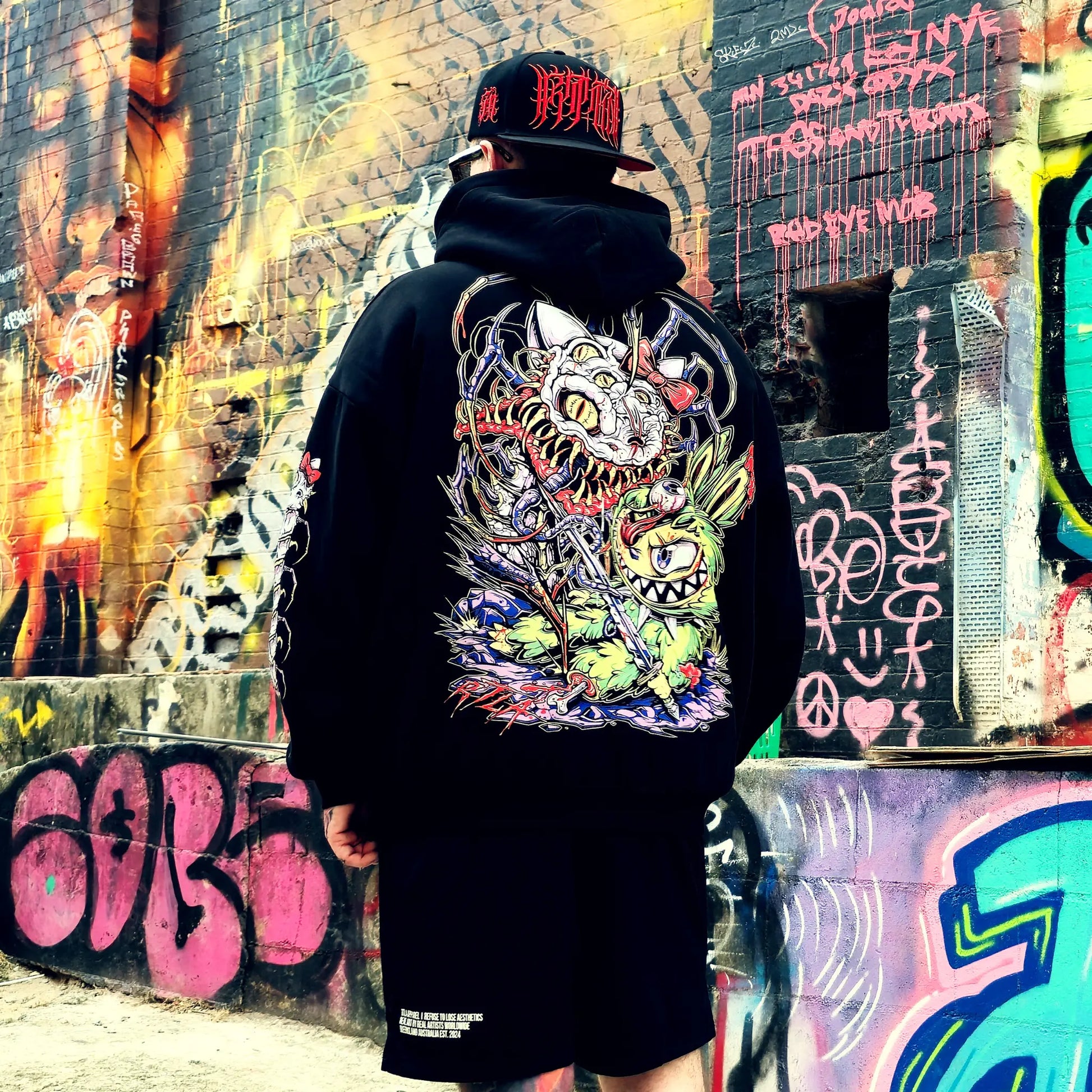 Person wearing a black hoodie with a colorful graphic design in front of a graffiti-covered wall.