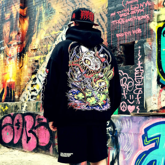 Person wearing a black hoodie with a colorful graphic design in front of a graffiti-covered wall.