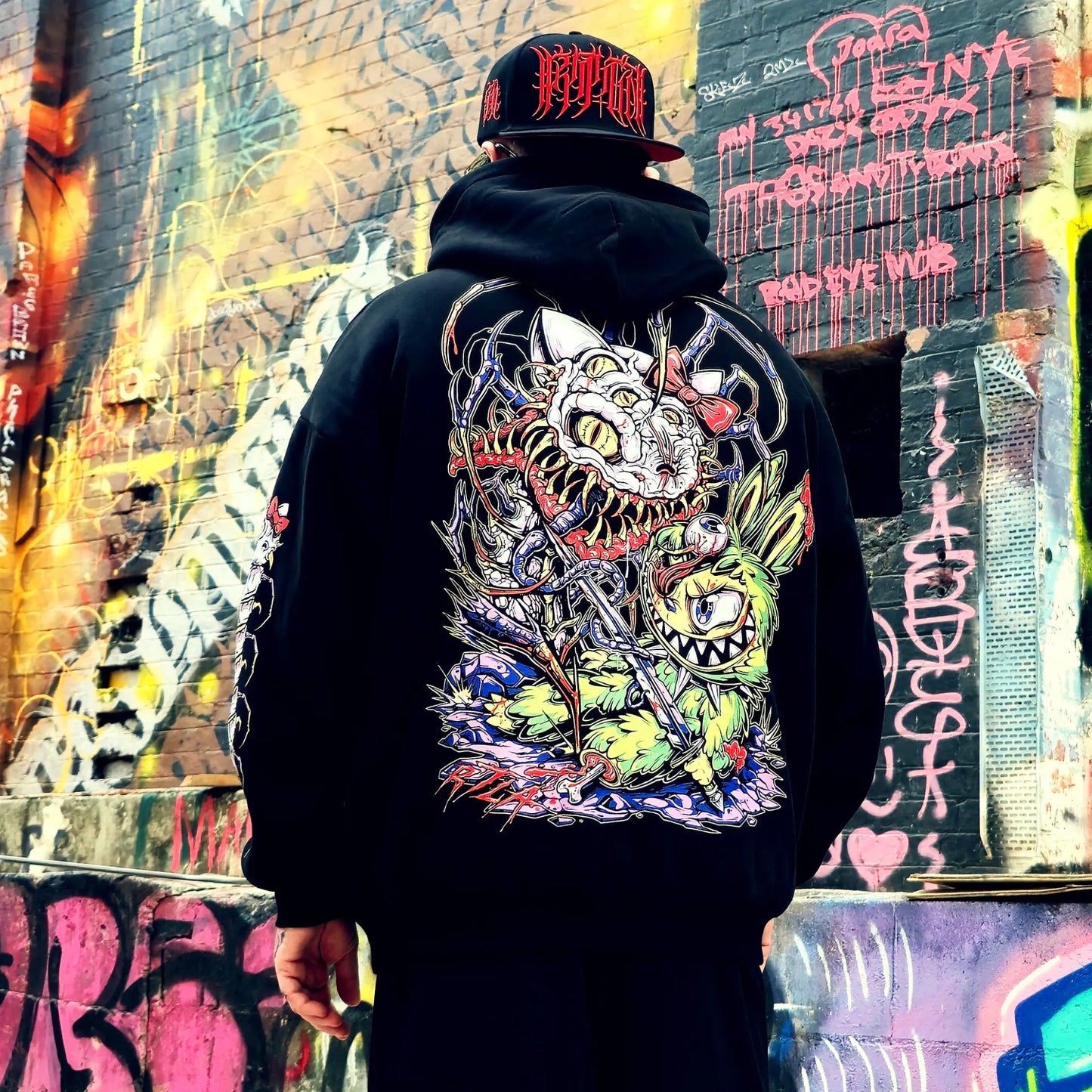 Person wearing a black hoodie with a colorful graphic design in front of a graffiti-covered wall.