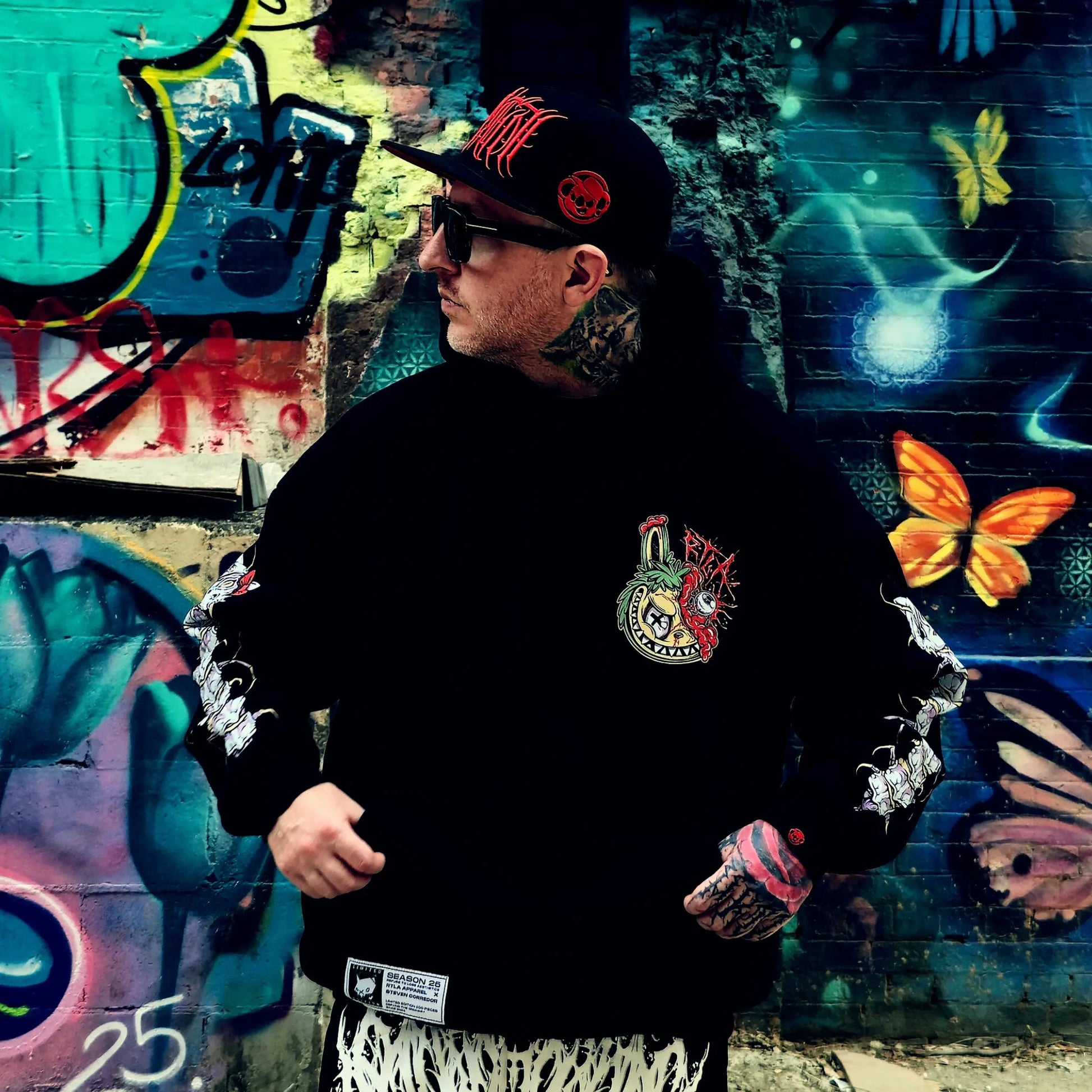 Person wearing a black hoodie with colorful designs in front of a graffiti wall.