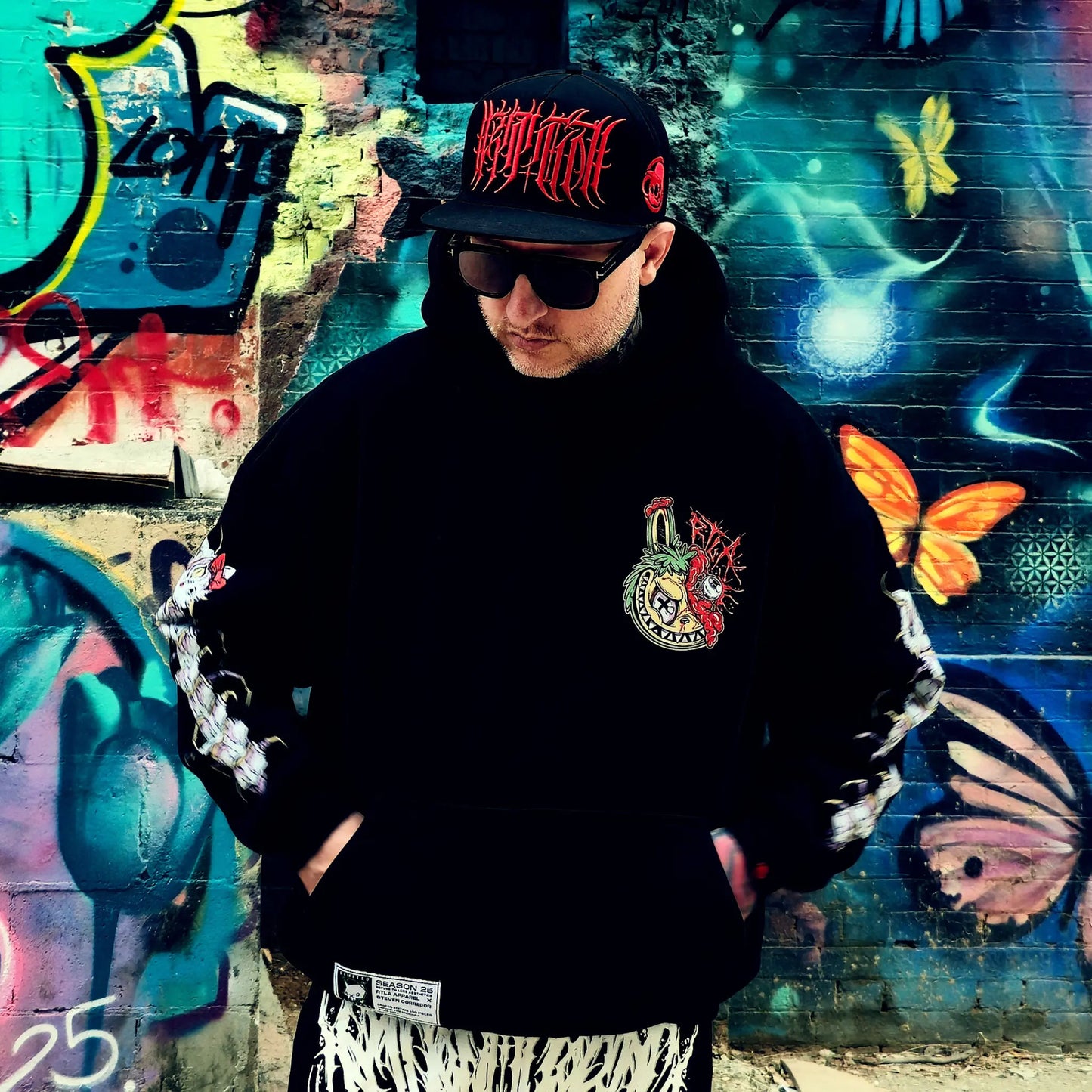 Person wearing a black hoodie with a colorful logo and a black cap in front of a graffiti wall.