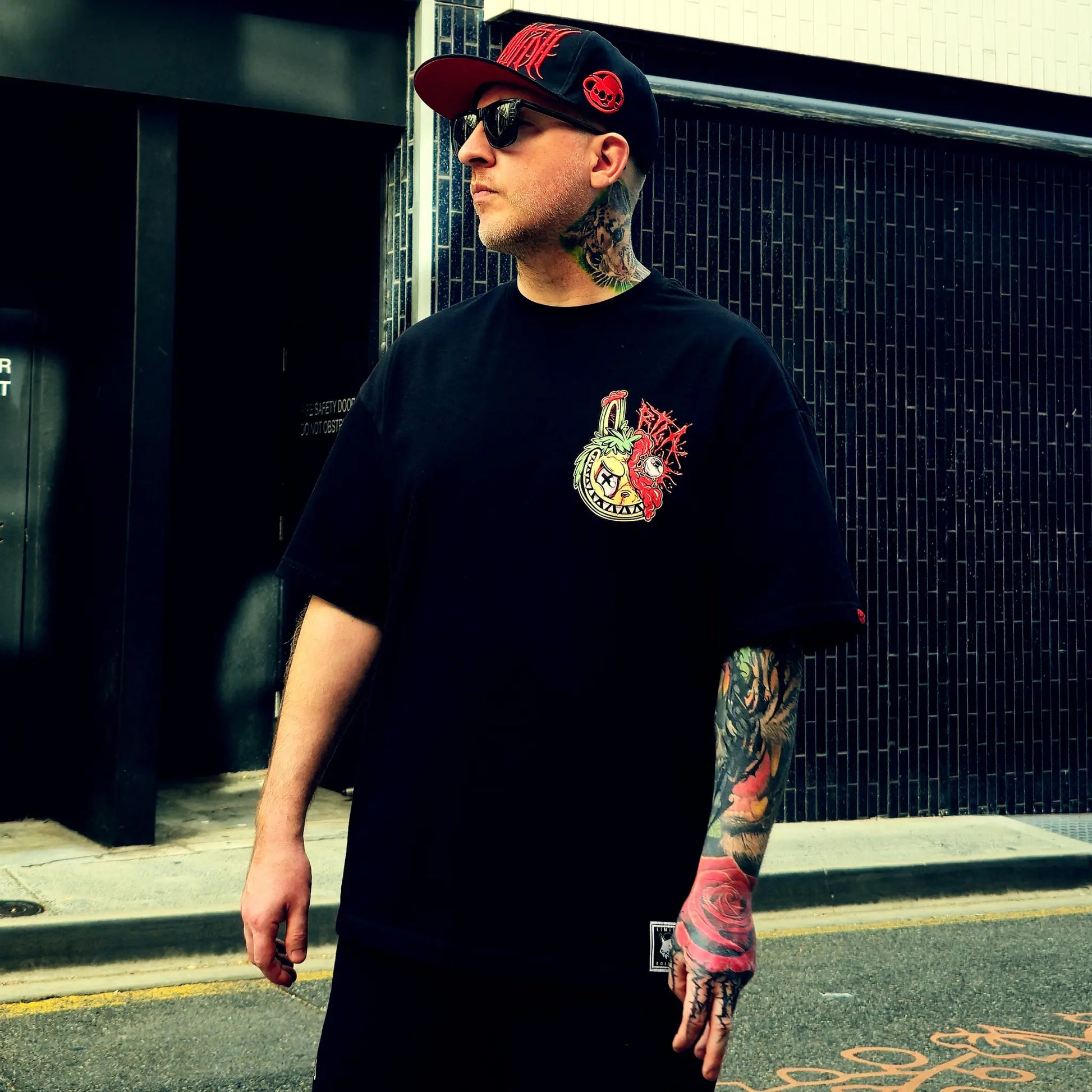 Man wearing a black t-shirt with a colorful logo and a cap, standing on a street.